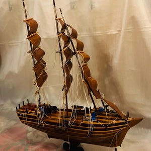 May include: A detailed wooden model of a sailing ship. The ship features multiple masts with brown sails and intricate rigging. The hull is a rich brown color, and the model sits on a small stand with an anchor detail. A decorative nautical item.