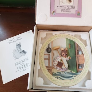以下が含まれることがあります： ビートリックス・ポターの「ふたりのわるいねずみ」の物語から、3Dイラストが描かれたセラミックプレート。ネズミはほうきとちりとりを持って、台所のシーンの中に立っています。プレートは装飾的な縁取りで囲まれており、薄い緑色の背景になっています。