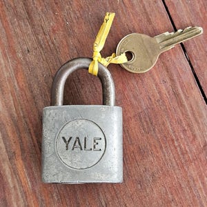 May include: A silver Yale padlock with a key and a yellow tag. The padlock has a rounded top and the word "YALE" in a circle on the front. The key is made of brass and has the words "MADE IN USA" on it.