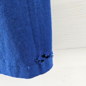 May include: Close-up of a section of a royal blue t-shirt. The bottom hem of the shirt has several holes and tears, indicating wear and tear. The fabric appears to be a soft knit material.