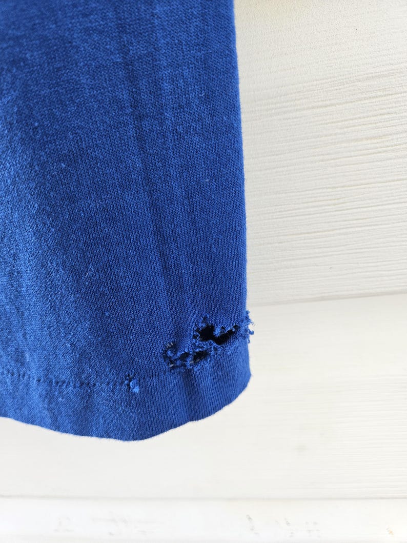 May include: Close-up of a section of a royal blue t-shirt. The bottom hem of the shirt has several holes and tears, indicating wear and tear. The fabric appears to be a soft knit material.