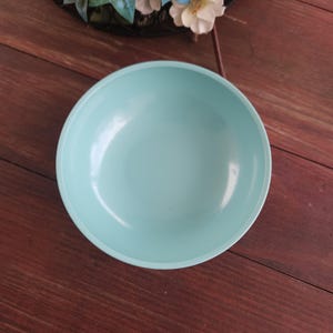 May include: A round, light blue bowl with a white rim. The bowl is empty and sits on a dark brown wooden surface. Decorative flowers and greenery are in the background.