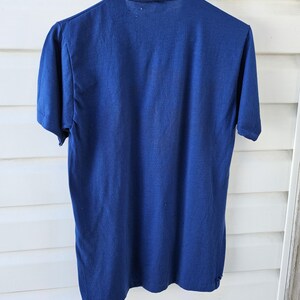 May include: A vintage navy blue short-sleeved t-shirt hangs from a metal hanger. The shirt's fabric appears worn, with visible wear and tear around the collar and hem. The background is a white wooden surface.