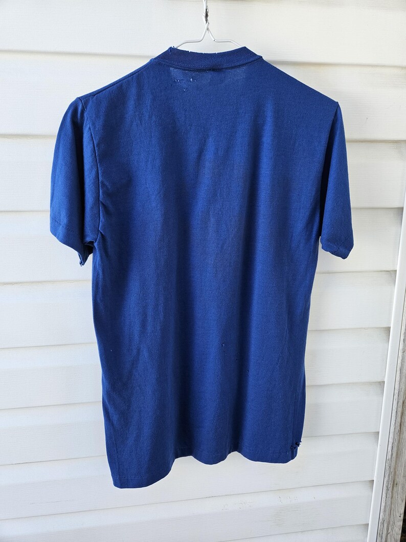 May include: A vintage navy blue short-sleeved t-shirt hangs from a metal hanger. The shirt's fabric appears worn, with visible wear and tear around the collar and hem. The background is a white wooden surface.