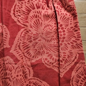 May include: Red fabric with a floral pattern. The fabric has a textured surface and a subtle sheen. The flowers are a lighter shade of red and have a detailed design.