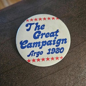 May include: A white button with a red and blue design. The button says "The Great Campaign Argo 1980" and has a ring of red stars around the edge.
