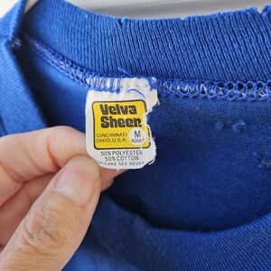 May include: Close-up of a blue t-shirt with a torn neckline. The tag reads "Velva Sheer" and includes the text "CINCINNATI OHIO, U.S.A. ADULT" and "50% POLYESTER 50% COTTON CARE SEE REVER".