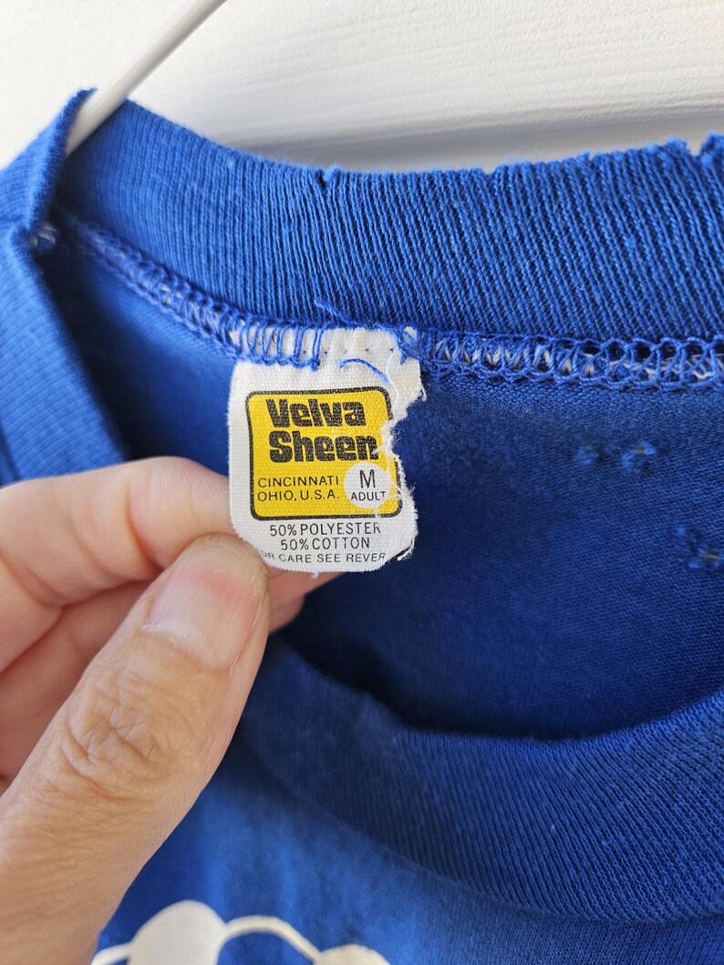 May include: Close-up of a blue t-shirt with a torn neckline. The tag reads "Velva Sheer" and includes the text "CINCINNATI OHIO, U.S.A. ADULT" and "50% POLYESTER 50% COTTON CARE SEE REVER".