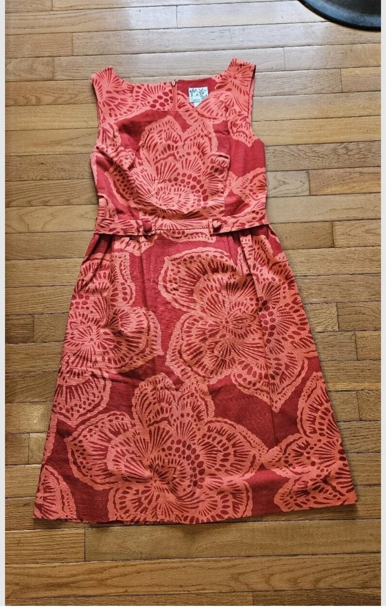 May include: A sleeveless red dress with a floral print. The dress has a fitted bodice and a full skirt. The dress is cinched at the waist with a belt.