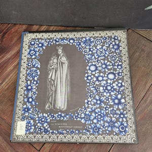 May include: A square vinyl record cover featuring a dark gray background with a central illustration of a crowned figure in a long robe. The border is decorated with blue and white floral patterns. Text reads Bellini La Straniera Montserrat Caballe.