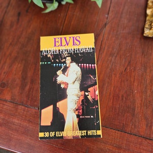 May include: A yellow and black VHS tape case with a picture of Elvis Presley on stage. The text on the case reads "Elvis Aloha From Hawaii 30 of Elvis Greatest Hits".