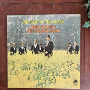 May include: A vintage vinyl record album cover featuring Herb Alpert & The Tijuana Brass. The cover art shows the band members standing in a field of yellow flowers. The album title "The Beat of the Brass" is printed in large, bold letters.
