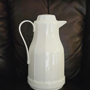 May include: A white, vintage thermos with a handle and a spout. The thermos has a geometric design with vertical ridges and a matching lid. The handle is curved, and the spout is small and angled. The thermos is made of a glossy material.