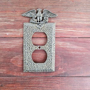 Puede incluir: Una cubierta decorativa de enchufe de estilo antiguo con una superficie texturizada y un emblema de águila. La cubierta tiene dos aberturas circulares y está hecha de un material metálico. El águila está posada sobre un escudo y ramas de olivo.