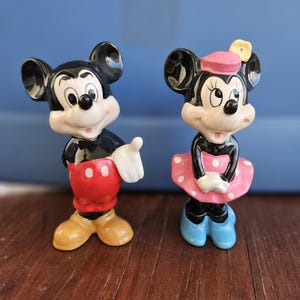 May include: A vintage ceramic figurine set of Mickey and Minnie Mouse. Mickey is wearing red shorts with white buttons and yellow shoes. Minnie is wearing a pink dress with white polka dots, a pink hat with a yellow flower, and blue shoes.
