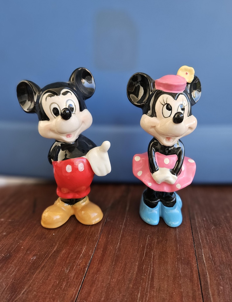 May include: A vintage ceramic figurine set of Mickey and Minnie Mouse. Mickey is wearing red shorts with white buttons and yellow shoes. Minnie is wearing a pink dress with white polka dots, a pink hat with a yellow flower, and blue shoes.