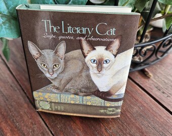 Op de afbeelding: Een bruin boek met de titel "The Literary Cat" met illustraties van twee katten. De omslag toont de tekst "Quips, quotes, and observations". De katten zijn afgebeeld op een stapel boeken.