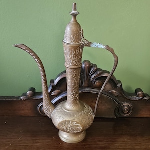 Puede incluir: Una jarra de agua antigua de latón con intrincados grabados florales. La jarra presenta un pico largo y curvo y un asa. El diseño incluye una base bulbosa y un cuello alto y esbelto, rematado con un adorno decorativo. La jarra es de color dorado cálido.