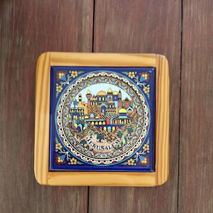 May include: A square wooden coaster featuring a central ceramic tile with a colorful illustration of the city of Jerusalem. The design includes a circular border with floral patterns and the text JERUSALEM printed at the bottom of the city scene.