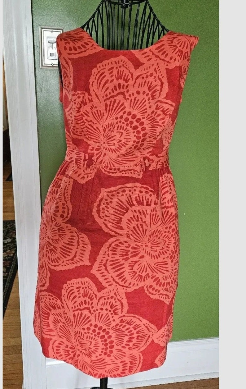 May include: A sleeveless red dress with a floral print. The dress has a fitted bodice and a flowing skirt. The flowers are a lighter shade of red and have a delicate, intricate design.