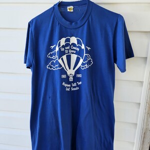 May include: A blue t-shirt with a white graphic of a hot air balloon, clouds, and text. The text reads "Up and Coming 20 Years" and "1963 1983 Keystone Tall Tree Girl Scouts."