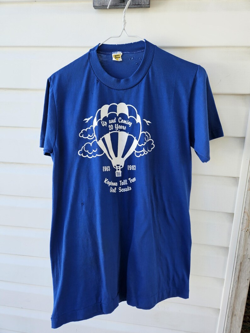 May include: A blue t-shirt with a white graphic of a hot air balloon, clouds, and text. The text reads "Up and Coming 20 Years" and "1963 1983 Keystone Tall Tree Girl Scouts."