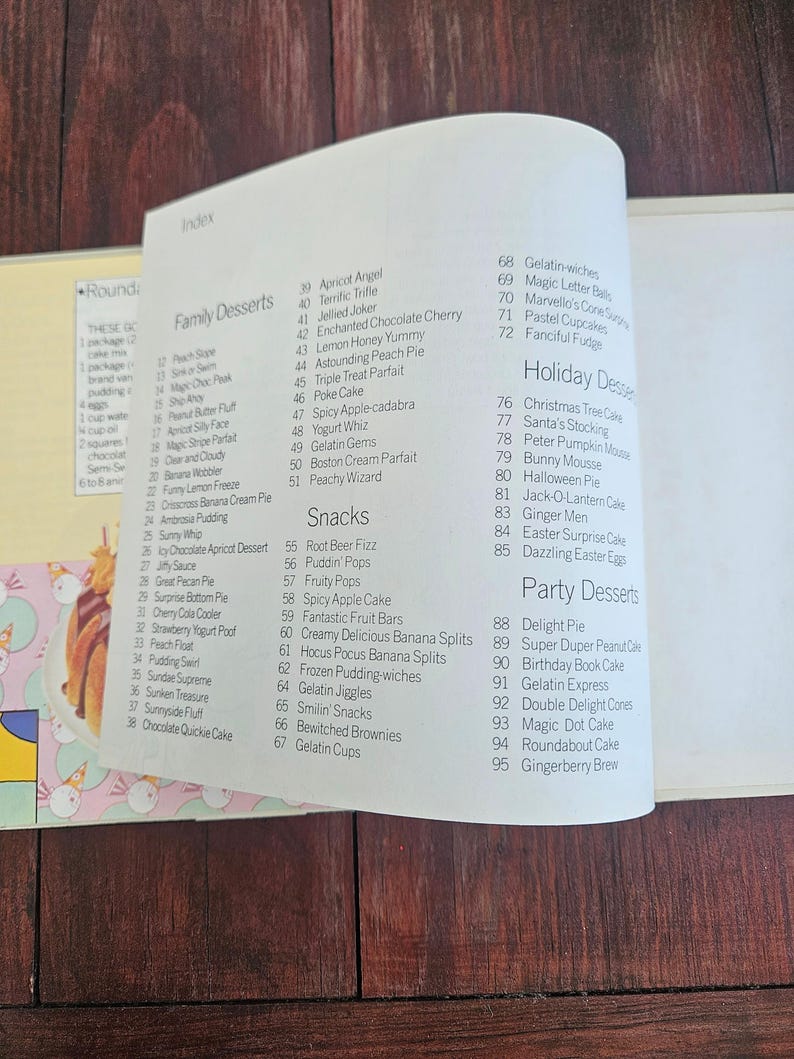 Op de afbeelding: Openstaande pagina uit een kookboek met dessertrecepten. De index bevat categorie&euml;n als Familiedesserts, Snacks en Feestdesserts. Titels zijn onder meer "Apricot Angel", "Gelatin Gems" en "Gingerberry Brew". Op een houten oppervlak.