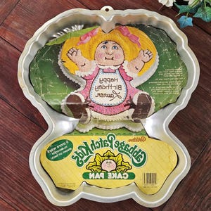 Puede incluir: Molde para pastel vintage de Cabbage Patch Kids. El molde tiene la forma de un Cabbage Patch Kid con cabello rubio y un vestido rosa. El molde tiene las palabras "Happy Birthday" y "XOXOX" en la parte delantera. El molde es de metal y tiene una etiqueta amarilla y verde.