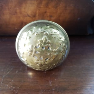 May include: A gold-colored metal knob with an ornate design. The knob has a circular shape and is decorated with a repeating pattern of leaves and flowers.