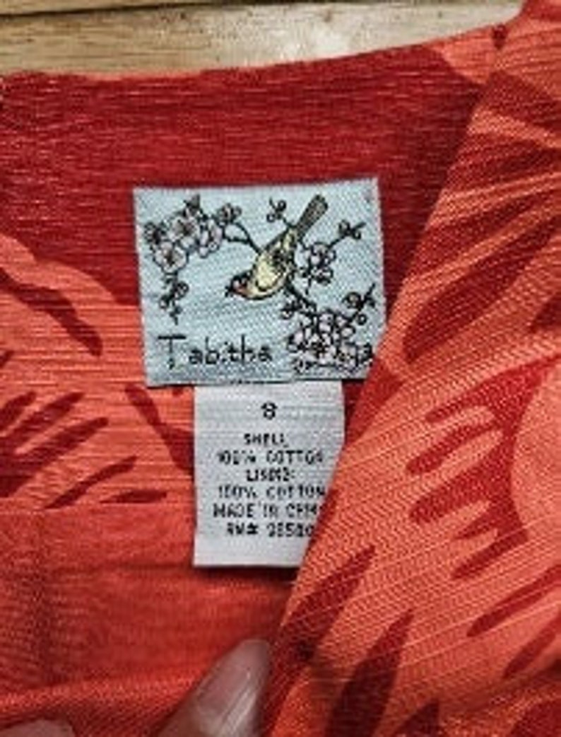 May include: A red and orange cotton dress with a floral print. The dress has a bird and flower design on the label. The label also says "Tabitha" and "8".