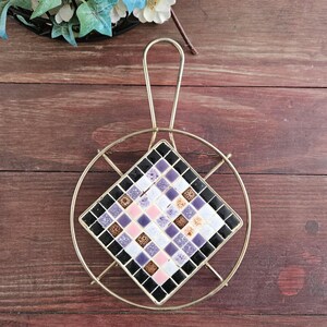 May include: A vintage trivet with a gold-toned metal frame and handle. The center features a diamond-shaped mosaic tile design in black, purple, pink, and white. The trivet is designed to protect surfaces from hot cookware.
