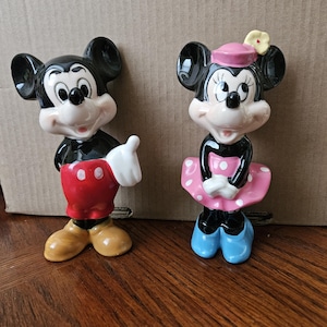 May include: A vintage ceramic figurine set of Mickey Mouse and Minnie Mouse. Mickey is wearing his signature red shorts and yellow shoes. Minnie is wearing a pink dress with white polka dots, a pink hat with a yellow flower, and blue shoes.