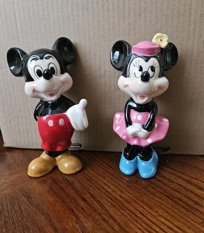 May include: A vintage ceramic figurine set of Mickey Mouse and Minnie Mouse. Mickey is wearing his signature red shorts and yellow shoes. Minnie is wearing a pink dress with white polka dots, a pink hat with a yellow flower, and blue shoes.