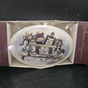 Puede incluir: Una caja de cerámica blanca ovalada con un diseño floral y una escena de un carruaje que lleva botellas de perfume. La caja tiene el texto "The Design Doesn't Wash Off" y "Laurie/Korgaard" en el lateral.