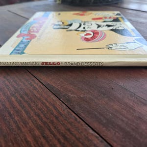 Op de afbeelding: Een vintage kookboek getiteld "Amazing Magical JELLO Brand Desserts" wordt getoond op een houten oppervlak. De omslag van het boek bevat kleurrijke illustraties en de rug toont de titel. Het boek is in goede staat.