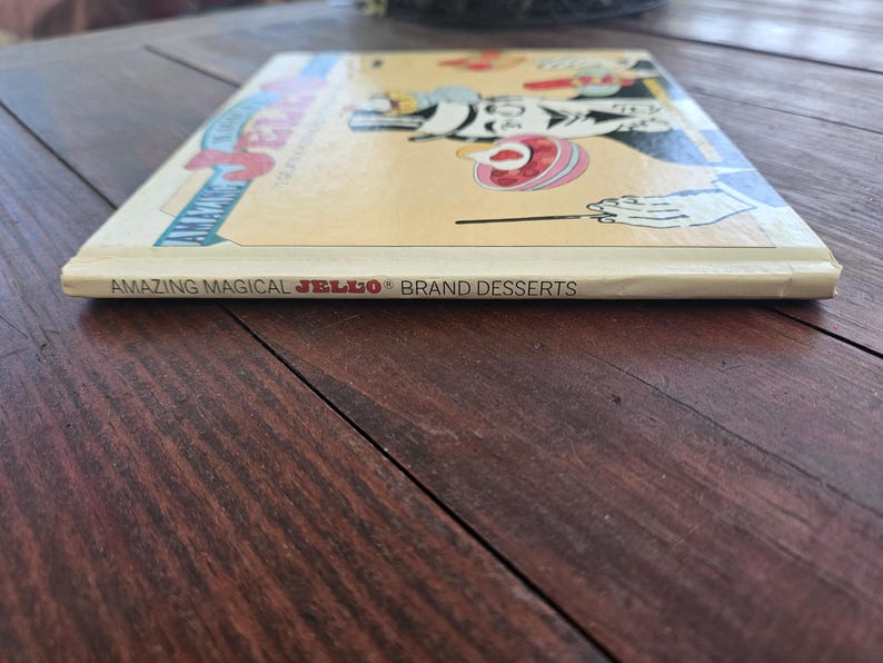 Op de afbeelding: Een vintage kookboek getiteld "Amazing Magical JELLO Brand Desserts" wordt getoond op een houten oppervlak. De omslag van het boek bevat kleurrijke illustraties en de rug toont de titel. Het boek is in goede staat.