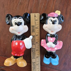 May include: A pair of ceramic Mickey Mouse and Minnie Mouse figurines. Mickey is wearing his signature red shorts and yellow shoes. Minnie is wearing a pink dress with white polka dots and blue shoes. Both figurines are approximately 6 inches tall.