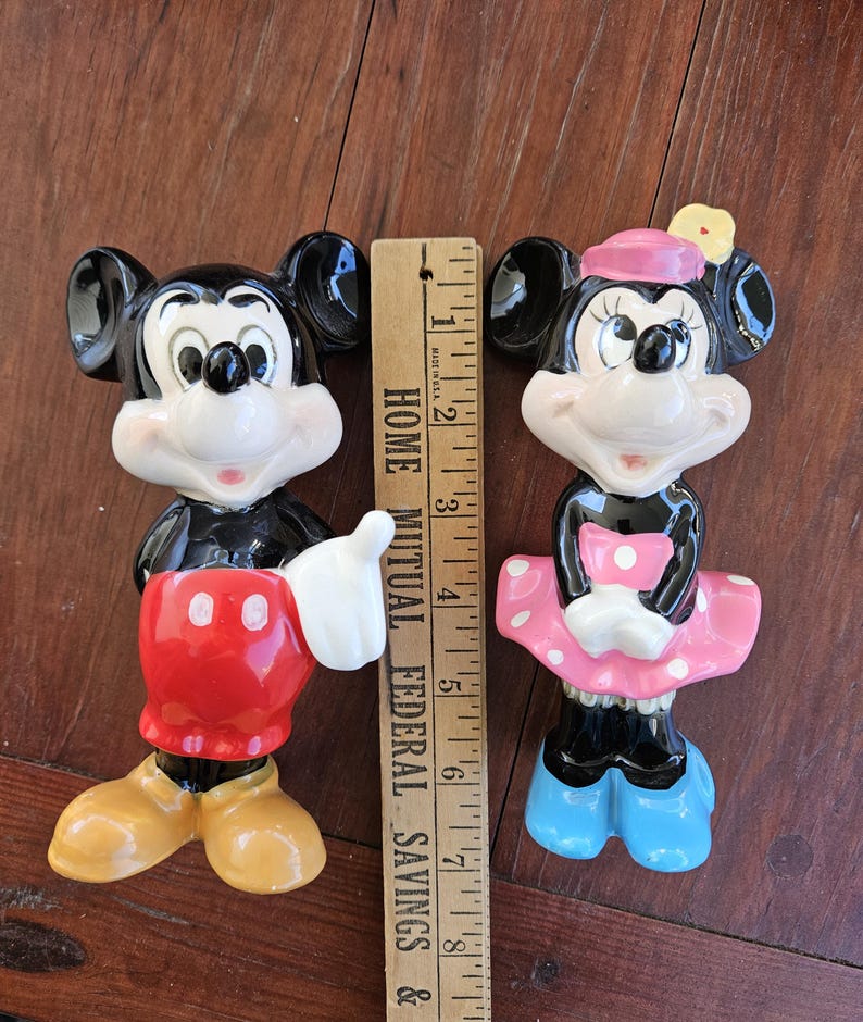 May include: A pair of ceramic Mickey Mouse and Minnie Mouse figurines. Mickey is wearing his signature red shorts and yellow shoes. Minnie is wearing a pink dress with white polka dots and blue shoes. Both figurines are approximately 6 inches tall.
