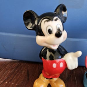 May include: A ceramic figurine of Mickey Mouse. Mickey is standing with his right arm outstretched and his left hand on his hip. He is wearing his signature red shorts with two white buttons and yellow shoes. His face is white with black eyes and a black nose. His ears are large and black.