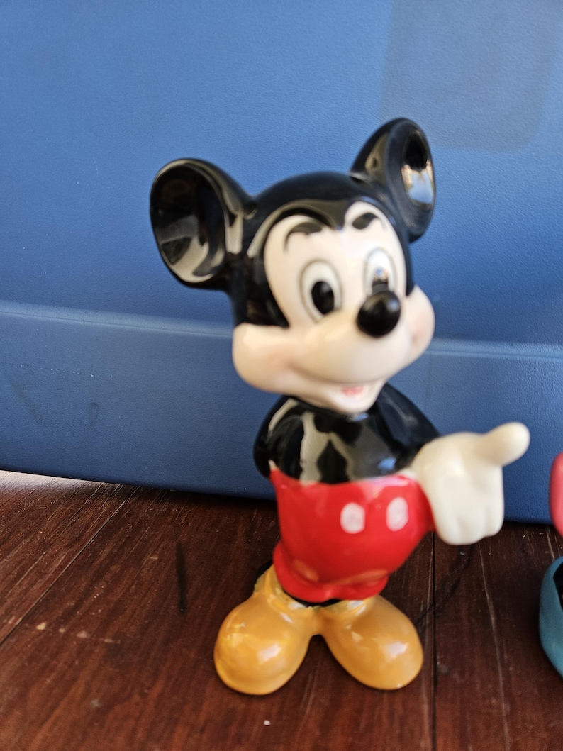 May include: A ceramic figurine of Mickey Mouse. Mickey is standing with his right arm outstretched and his left hand on his hip. He is wearing his signature red shorts with two white buttons and yellow shoes. His face is white with black eyes and a black nose. His ears are large and black.