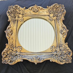 May include: A gold ornate mirror with a round glass inset. The frame has a detailed, intricate design with a distressed finish.