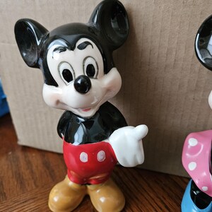 May include: A ceramic figurine of Mickey Mouse, standing with his right hand raised. He is wearing his signature red shorts with two white buttons, black shoes, and white gloves. His ears are black, and his face is white with black eyes and a black nose.
