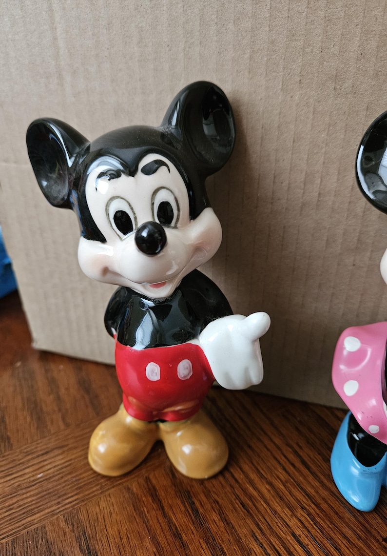 May include: A ceramic figurine of Mickey Mouse, standing with his right hand raised. He is wearing his signature red shorts with two white buttons, black shoes, and white gloves. His ears are black, and his face is white with black eyes and a black nose.