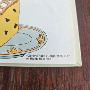 Op de afbeelding: Een vintage illustratie van een stuk gele cake met donkere chocoladeschilfers op een lichtblauw bord met gouden rand. De afbeelding bevat de tekst "General Foods Corporation 1977 All Rights Reserved."