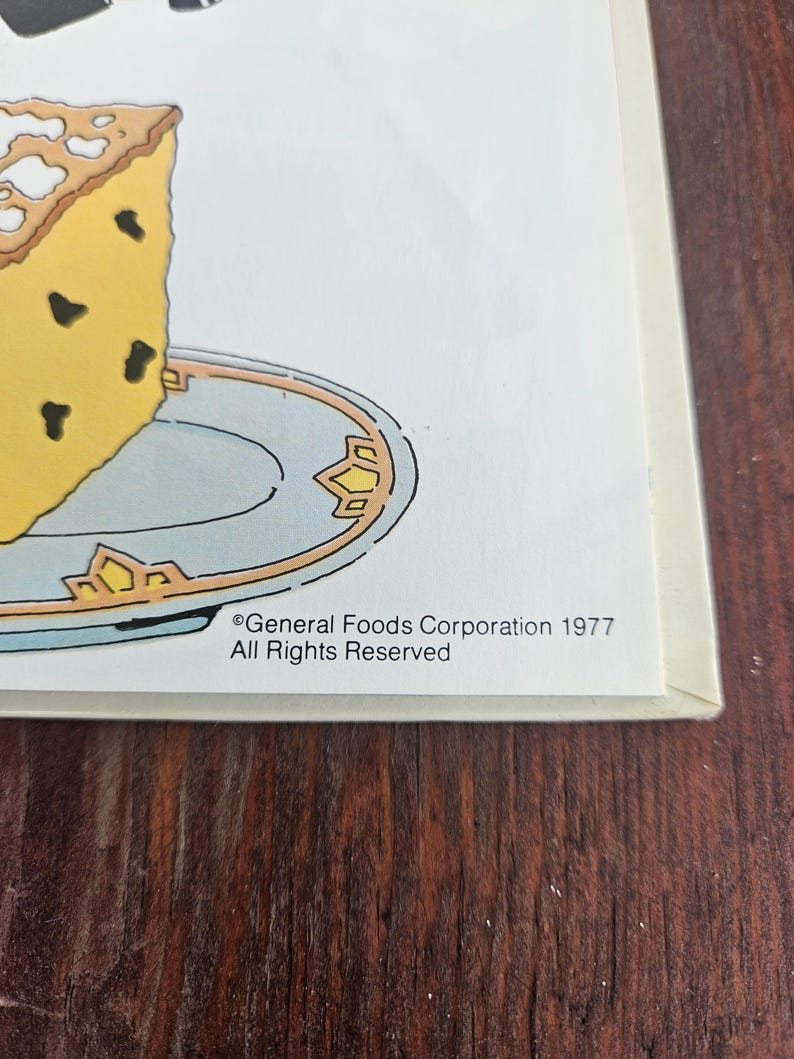 Op de afbeelding: Een vintage illustratie van een stuk gele cake met donkere chocoladeschilfers op een lichtblauw bord met gouden rand. De afbeelding bevat de tekst "General Foods Corporation 1977 All Rights Reserved."