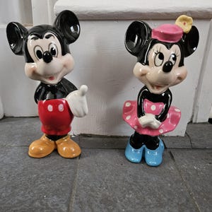 May include: A pair of ceramic figurines of Mickey Mouse and Minnie Mouse. Mickey is wearing red shorts with white polka dots and yellow shoes. Minnie is wearing a pink dress with white polka dots, a pink hat with a yellow flower, and blue shoes.