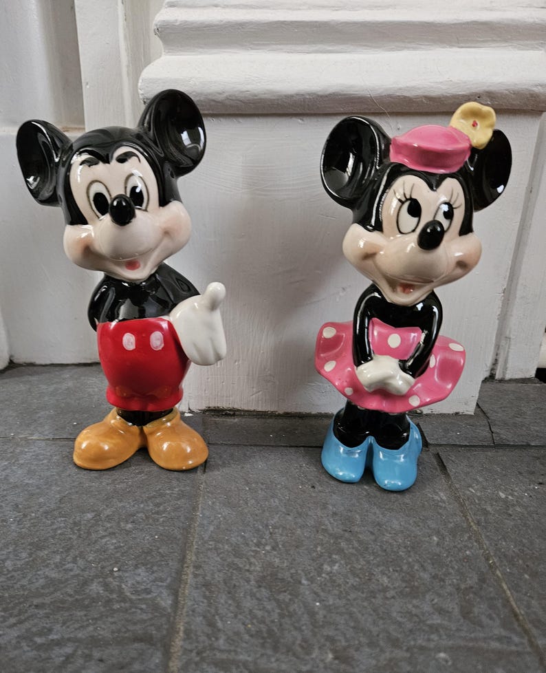 May include: A pair of ceramic figurines of Mickey Mouse and Minnie Mouse. Mickey is wearing red shorts with white polka dots and yellow shoes. Minnie is wearing a pink dress with white polka dots, a pink hat with a yellow flower, and blue shoes.