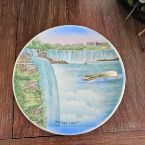 May include: A round, decorative plate featuring a watercolor painting of Niagara Falls. The plate has a blue and white border, with the words "Niagara Falls" at the bottom. The image depicts the falls with a boat and buildings in the background.