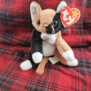 Puede incluir: Un peluche Beanie Baby de gato calico negro, blanco y marrón con una etiqueta Ty roja y blanca que dice "Beanie Babies Baby".
