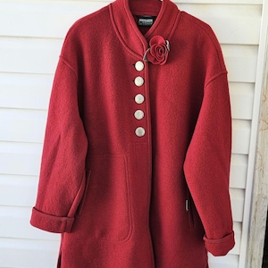 May include: A red wool coat with a round collar and large silver buttons. The coat has a rose-shaped pin on the collar and cuffed sleeves. The coat is hanging on a white hanger against a white wooden background.